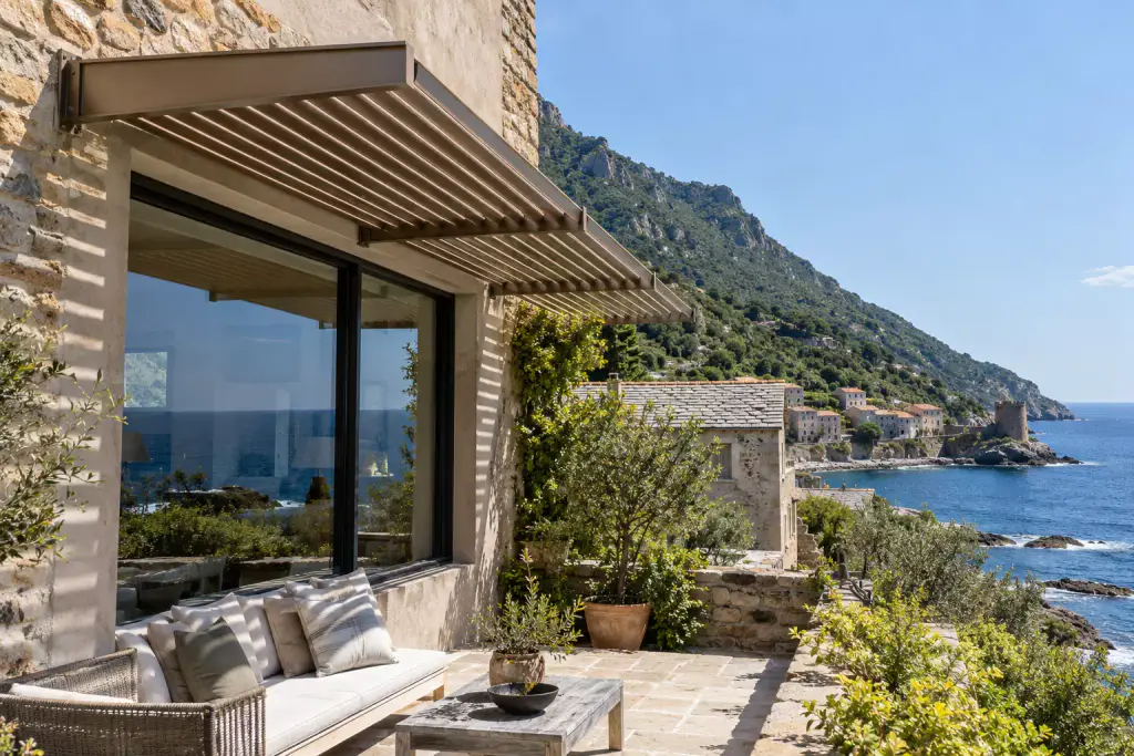 Brise-soleil à lames d’aluminium bronze posé en casquette au-dessus d’une grande baie vitrée, dans un mur de pierre ; l’ombre rayée des lames sur la façade et la banquette blanche du salon. À droite, un toit de lauzes, un village au bord de l’eau avec une tour ronde, et une côte abrupte couverte de maquis.