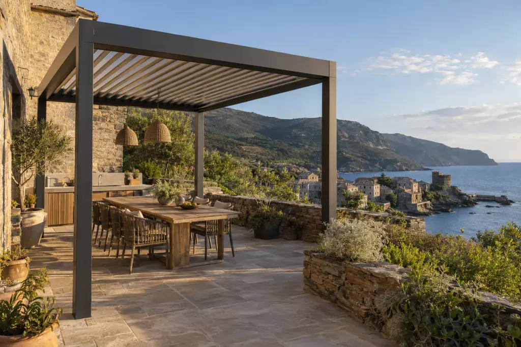 Pergola à structure anthracite et lames ouvertes, devant le mur d’une maison de pierre ; dessous, une longue table en bois, des chaises tressées, deux suspensions en osier et une cuisine d’été. Au-delà du muret de pierre sèche, un village au bord de l’eau, une tour ronde sur la pointe et la côte montagneuse.