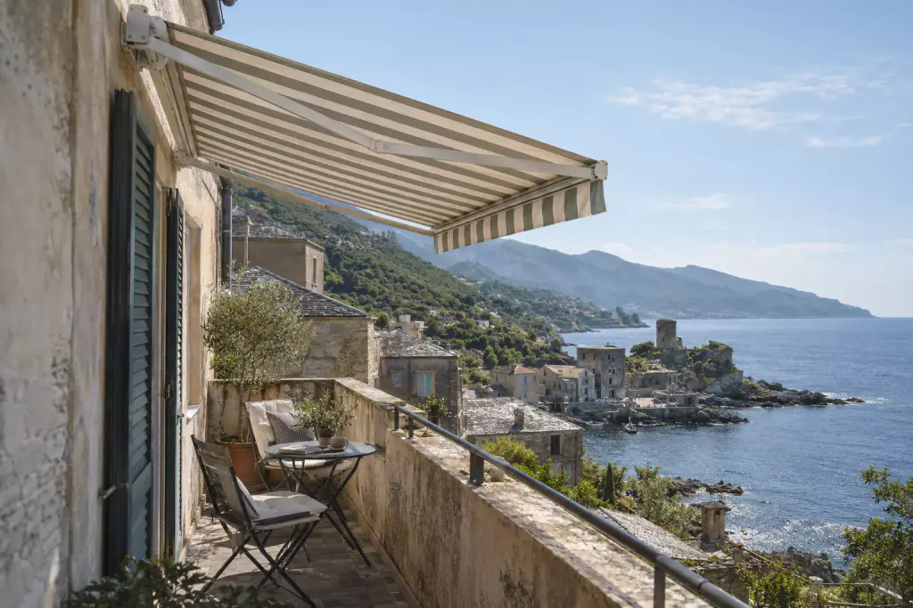 Store banne à rayures vertes et crème déployé au-dessus d’un étroit balcon de maison ancienne, volets verts, petite table de bistrot et deux chaises pliantes ; en contrebas, des toits de lauzes, un village au bord de l’eau avec une tour sur la pointe, et la côte montagneuse.