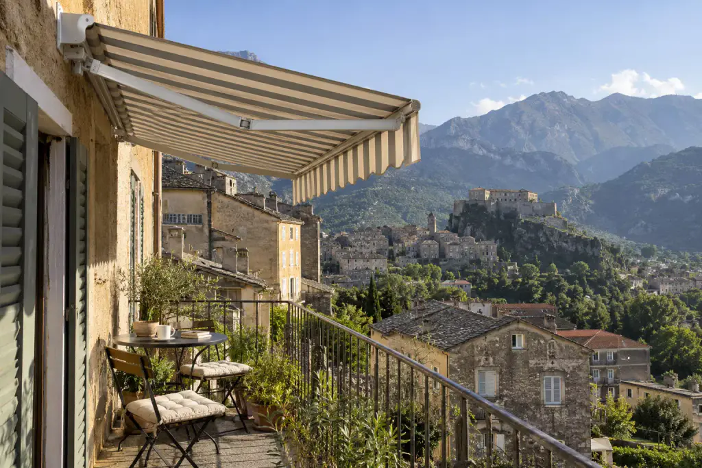 Store banne à rayures beiges et grises déployé au-dessus d’un balcon étroit, sur la façade d’une vieille maison aux volets verts : table de bistrot, chaises à coussins, plantes en pot ; en contrebas, des toits de pierre, une ville perchée avec sa citadelle sur un rocher, des montagnes.