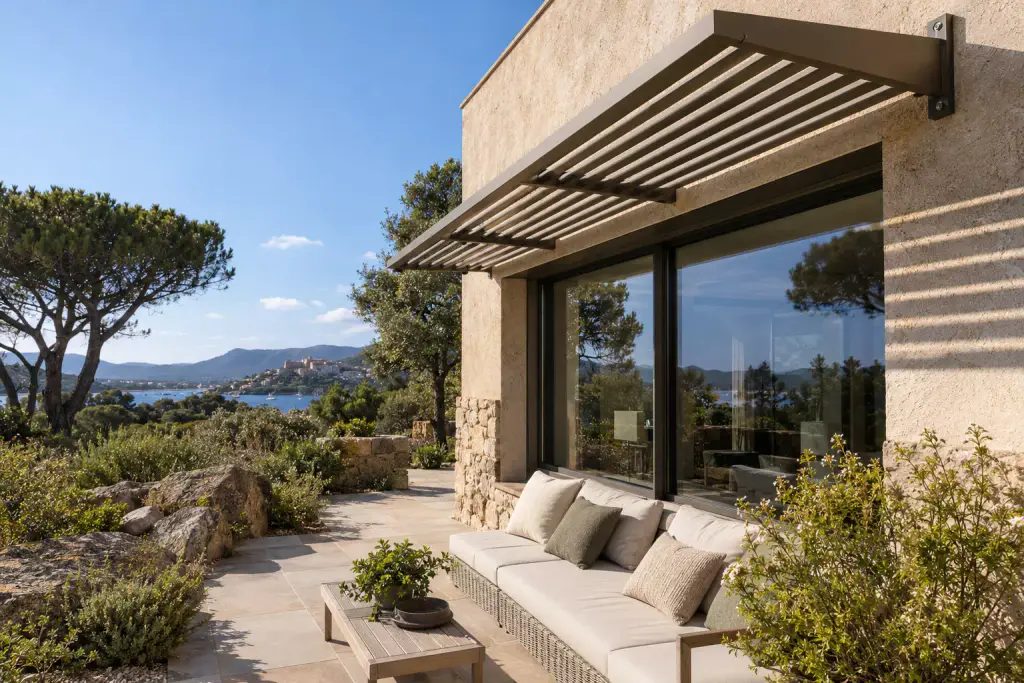 Brise-soleil à lames d’aluminium bronze posé en casquette au-dessus d’une grande baie vitrée, sur une façade enduite et en pierre ; un canapé blanc, une table basse en bois, des rochers de granit, des pins et un golfe au loin.