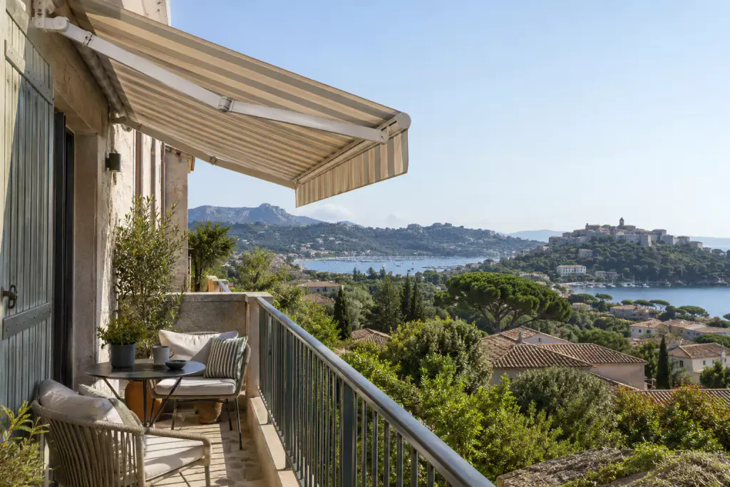Store banne à rayures beige et vert tendre déployé au-dessus d’un balcon en pierre : deux fauteuils, une table ronde, un garde-corps en métal ; au loin, un golfe, un port de plaisance et une vieille ville sur sa colline.