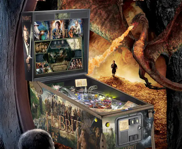 Le flipper The Hobbit, sa caisse décorée et son fronton, posé devant une grande image du film où un dragon souffle du feu.