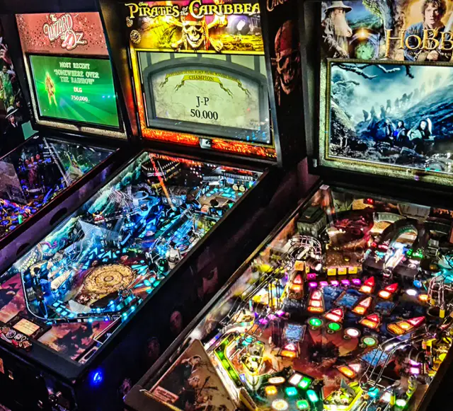 Trois flippers Jersey Jack Pinball allumés côte à côte, vus d’en haut : frontons à écran, plateaux éclairés de couleurs.