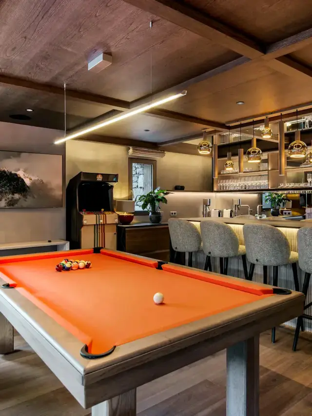 Une salle de jeux au plafond de bois à caissons : un billard Maison Toulet à drap orange au premier plan, un bar et ses tabourets, des étagères éclairées au fond.