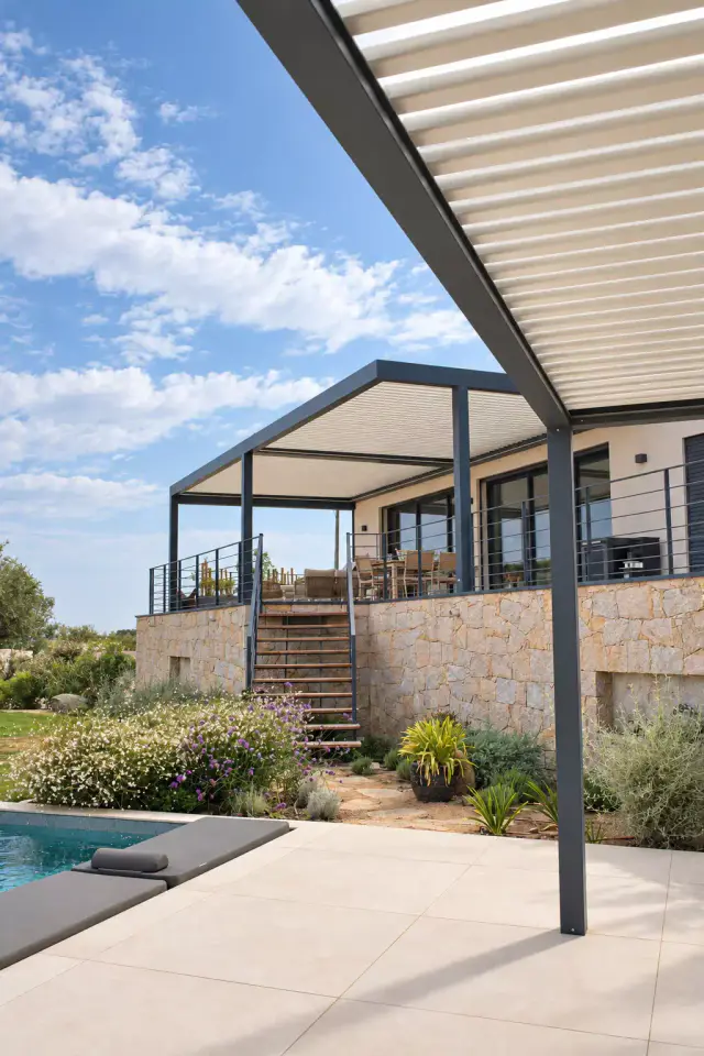 Deux pergolas bioclimatiques gris anthracite : l’une au premier plan vue par-dessous, l’autre au fond sur la terrasse haute de la villa, au-dessus d’un jardin de rocaille.