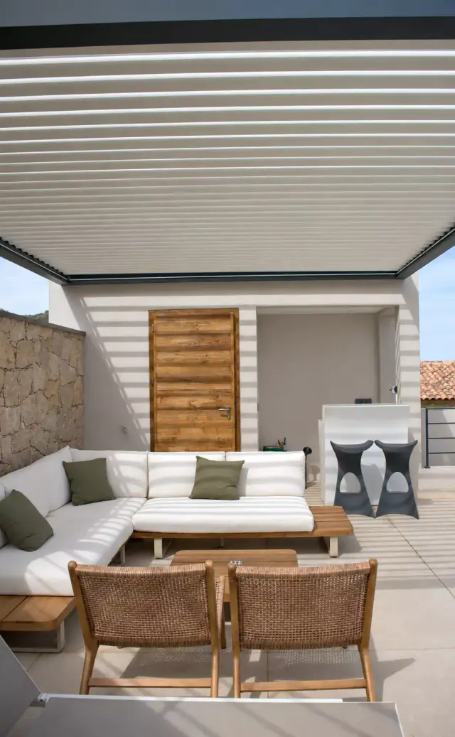 Pergola bioclimatique aux lames claires entrouvertes au-dessus d’un canapé d’angle blanc aux coussins verts et de deux fauteuils en bois tressé, devant une porte en bois.