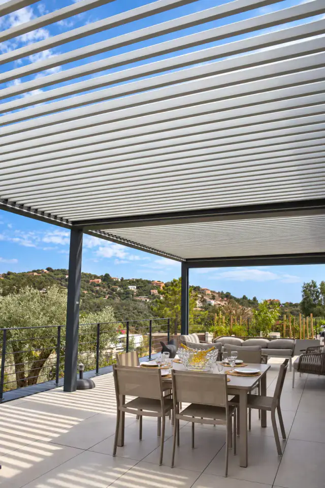 Sous une pergola bioclimatique aux lames claires entrouvertes, une table dressée et ses chaises taupe sur une terrasse carrelée, ombres rayées au sol, garde-corps, oliviers et collines boisées sous le ciel bleu.