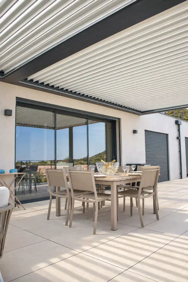 Pergola bioclimatique aux lames claires entrouvertes au-dessus d’une table à manger et de ses chaises taupe, sur une terrasse carrelée clair devant une baie vitrée coulissante.