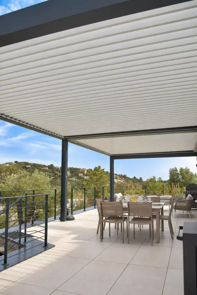 Pergola bioclimatique aux lames claires fermées formant un plafond continu au-dessus d’une table à manger taupe, terrasse ouverte sur des collines boisées.
