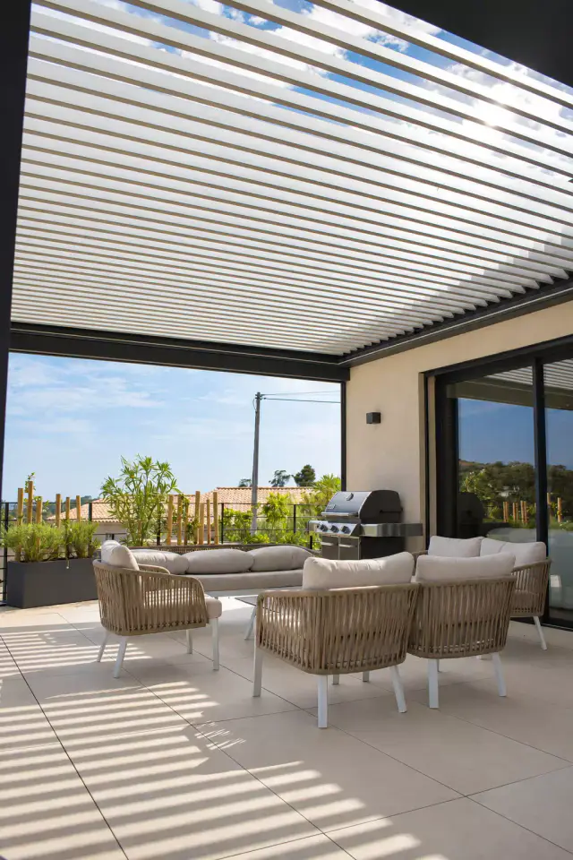 Pergola bioclimatique aux lames entrouvertes dessinant des ombres rayées sur le carrelage, au-dessus d’un salon d’extérieur en corde tressée beige et d’un barbecue à gaz.