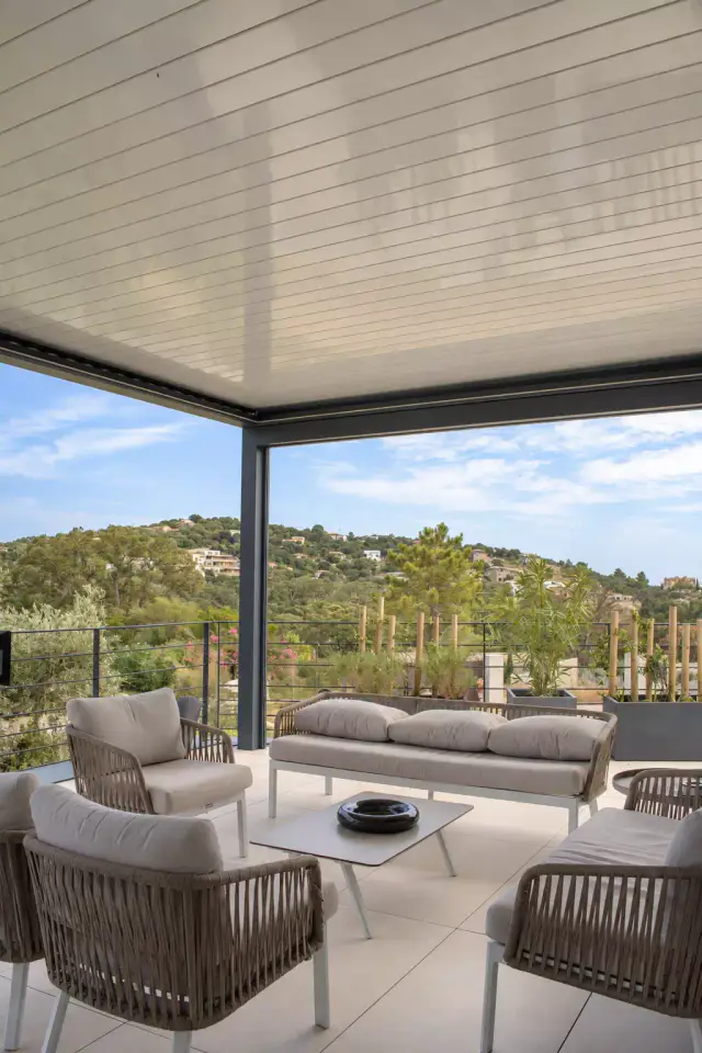 Pergola bioclimatique aux lames claires fermées abritant un salon d’extérieur en corde tressée et coussins beiges, autour d’une table basse, face aux collines boisées.