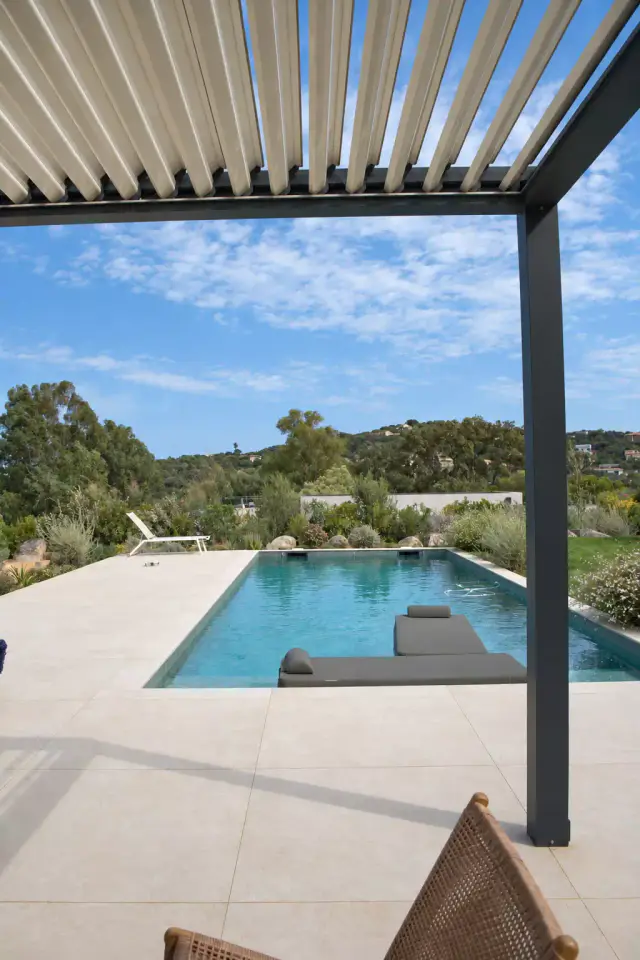 Pergola bioclimatique aux lames ouvertes encadrant la vue sur une piscine tout en longueur, sa plage carrelée clair et un jardin méditerranéen sous un ciel bleu.