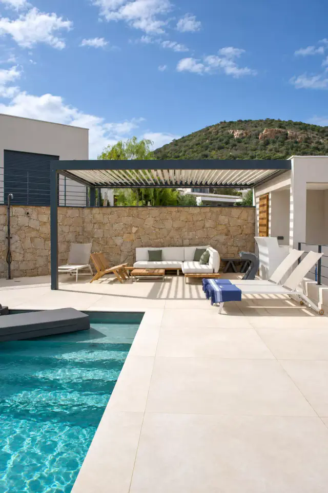 Piscine au premier plan et plage carrelée clair menant à une pergola bioclimatique gris anthracite adossée à un mur de pierre, bains de soleil et colline boisée au fond.