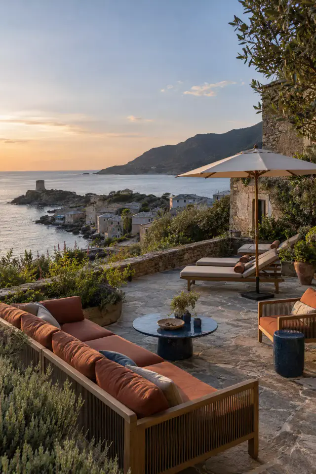 Terrasse dallée de pierre au coucher du soleil : canapé en bois aux coussins terracotta, table basse ronde bleue, fauteuil, bains de soleil sous un parasol à mât de bois ; au-delà d’un muret, un village au bord de l’eau, une tour sur la pointe et la mer.