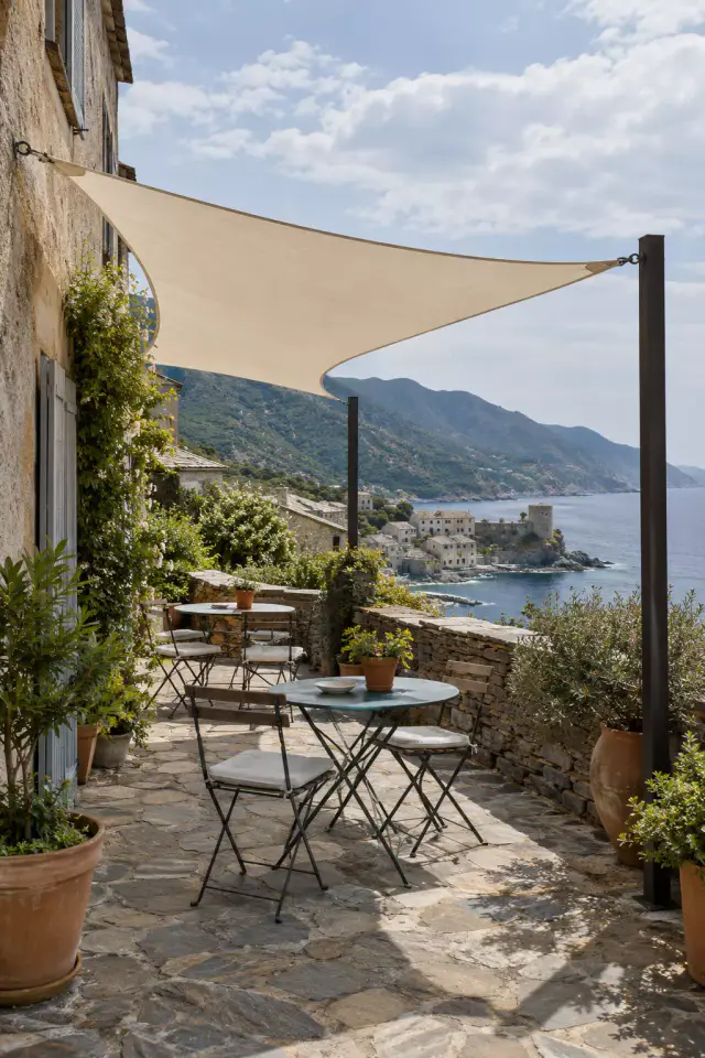 Voile d’ombrage triangulaire couleur sable, accrochée au mur d’une maison de pierre et tendue sur deux mâts noirs, au-dessus d’une terrasse dallée de pierre avec deux tables de bistrot et des pots d’olivier ; au-delà du muret, un village au bord de l’eau avec une tour, et la côte.