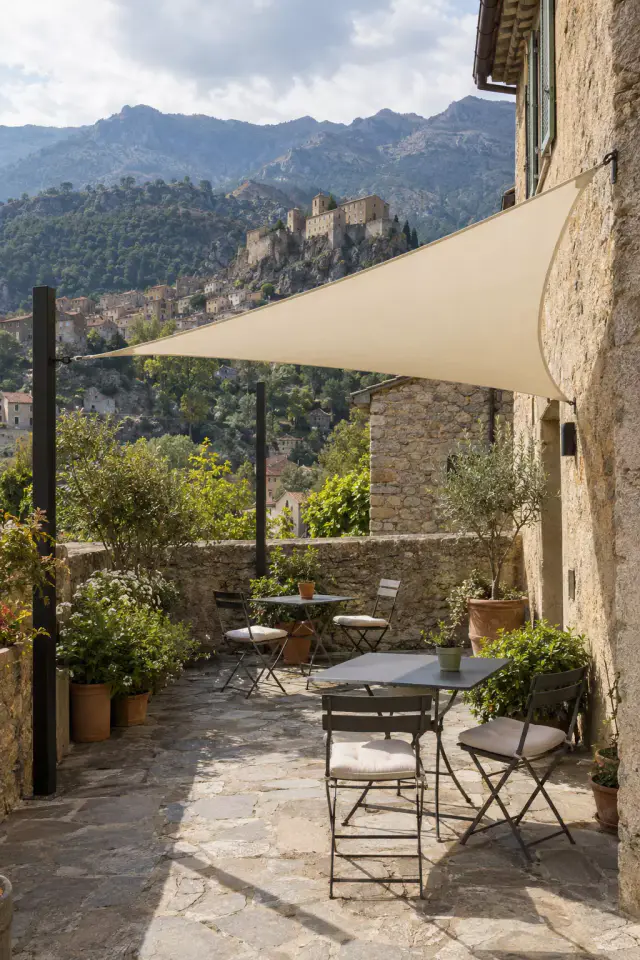 Voile d’ombrage triangulaire couleur crème tendue entre le mur d’une maison en pierre et un mât noir, au-dessus d’une terrasse dallée : tables et chaises pliantes de bistrot, plantes en pot, muret de pierre ; au fond, une citadelle sur son rocher, des maisons et des montagnes.