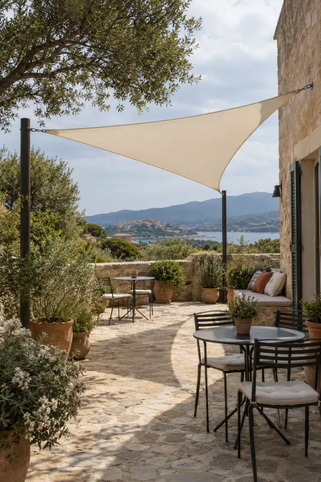 Voile d’ombrage triangulaire couleur crème tendue entre deux mâts noirs et le mur en pierre d’une maison, au-dessus d’une terrasse dallée de pierre : tables de bistrot, banquette à coussins, oliviers en pot ; un golfe et une ville sur sa colline au loin.
