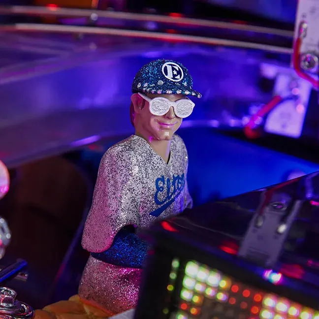 Sur le plateau du flipper Elton John, la figurine du chanteur en veste pailletée et casquette, assise au piano.