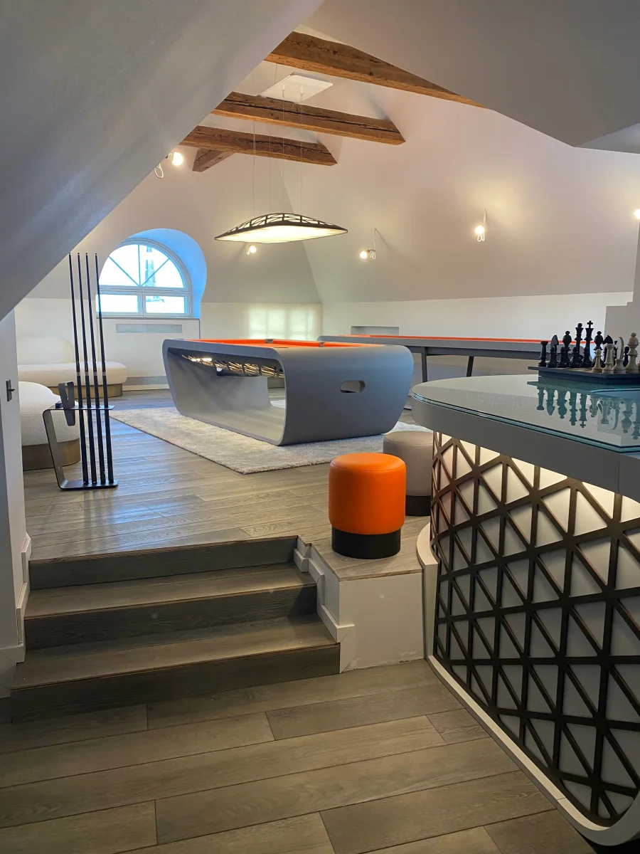 Une salle de jeux sous les combles, poutres apparentes et fenêtre en demi-lune : un billard gris à drap orange sur un tapis clair, un râtelier de queues, un pouf orange et, au premier plan, un bar à jeu d’échecs.