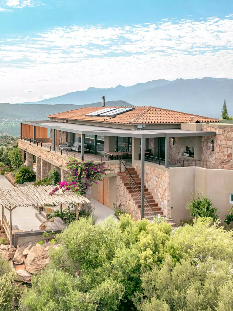 Drone view: stone villa with a tiled roof clinging to the slope, outdoor staircase, bougainvillea and wooden trellis above the rocks, maquis in the foreground, mountains beyond.
