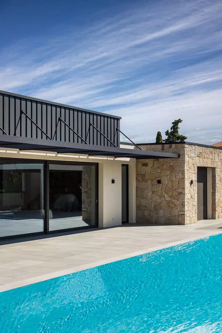 Une villa de pierre et de bardage noir sous une casquette de lames tenue par des tirants, vue depuis la piscine.