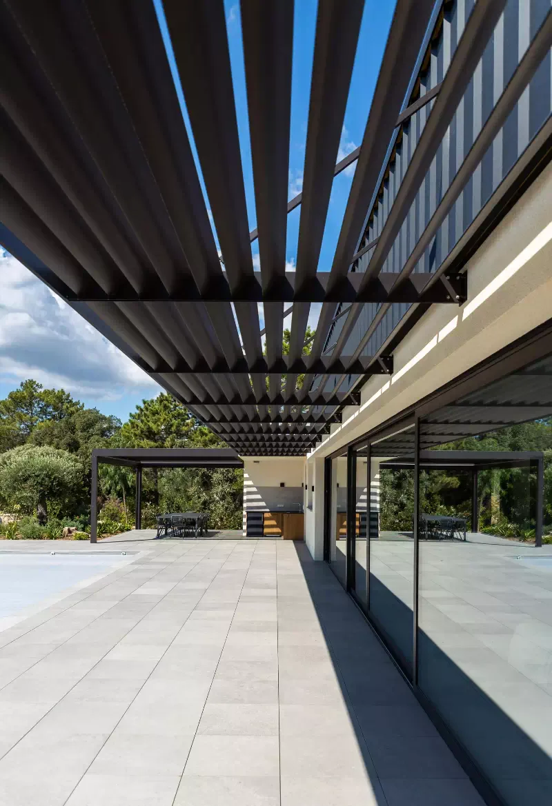 Anthracite louvred brise-soleil canopy seen from below, along the glazed façade; pale grey paving, the summer kitchen pergola in the background in front of the pines.