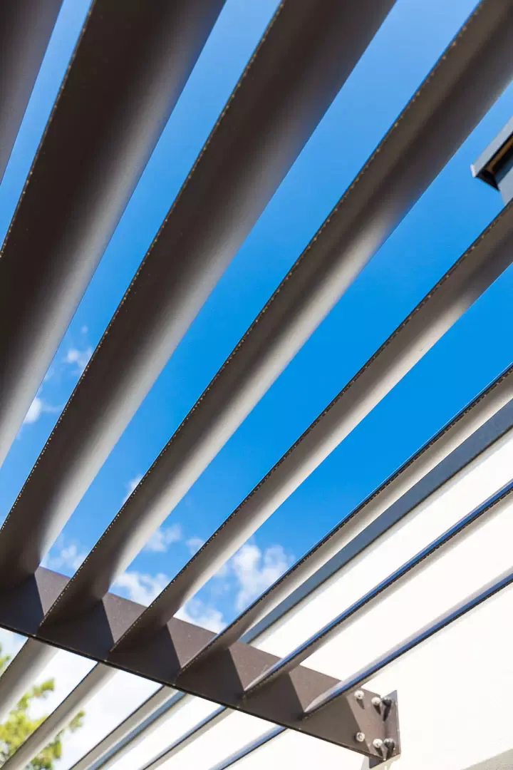 Lames du brise-soleil vues en contre-plongée et à contre-jour : profils gris en éventail sur le ciel bleu, platine de fixation et tirant en bas de l’image.