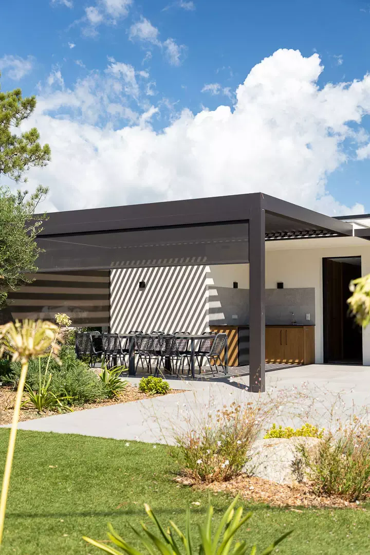 Anthracite bioclimatic pergola sheltering the summer kitchen and its table, side blind half lowered striping the white wall with shadow, garden in the foreground, cumulus clouds.