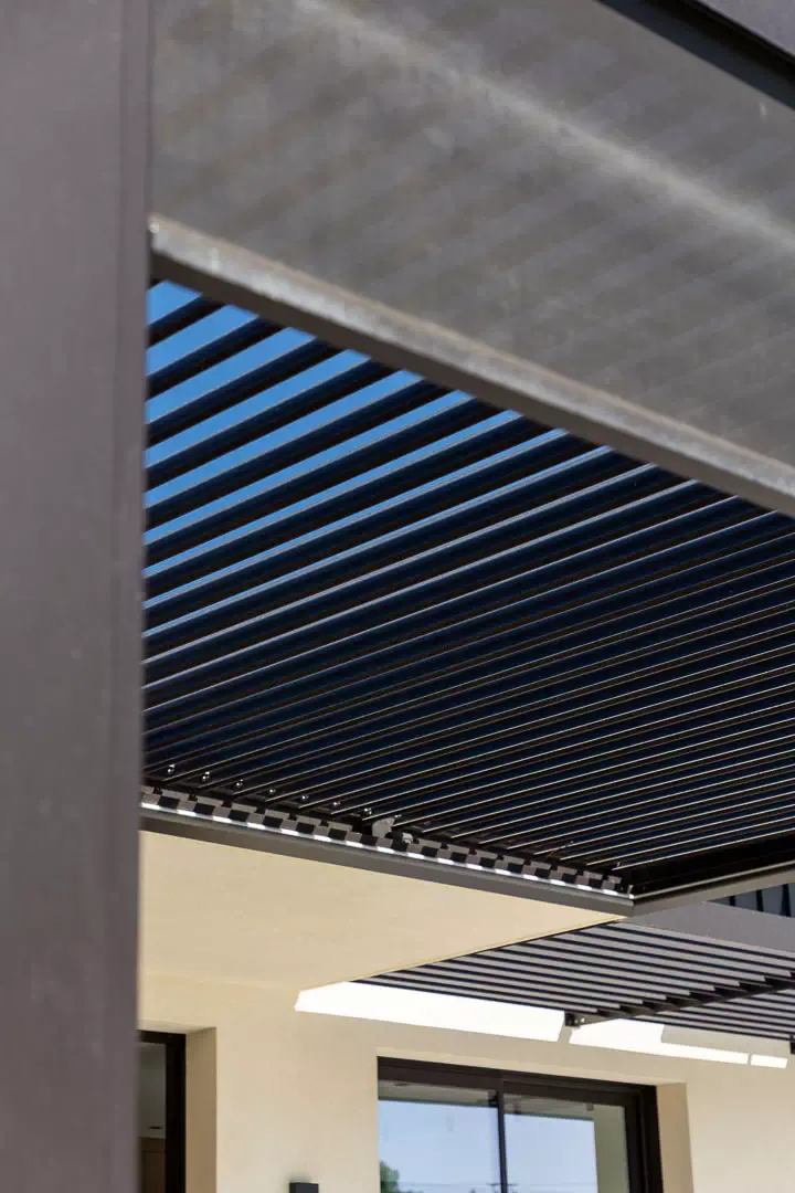 Lames anthracite de la pergola bioclimatique entrouvertes sur le ciel bleu, vues de dessous entre un poteau et la toile d’un store ; casquette brise-soleil et façade claire au fond.