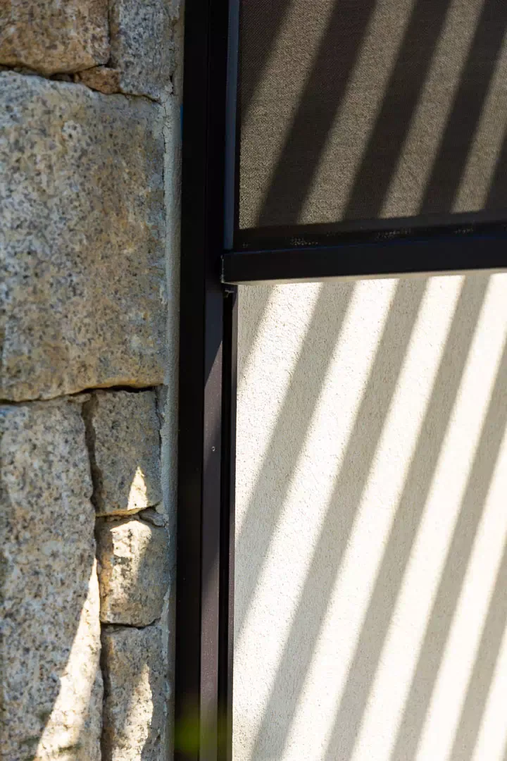 Bas d’un store screen à toile micro-perforée sombre et sa barre de charge dans la coulisse anthracite du poteau, mur de granit à gauche, enduit blanc rayé d’ombres dessous.